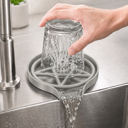 Hand using a grey press-type glass rinser on a stainless steel sink deck, with water spraying inside an upside-down glass and draining into the sink.